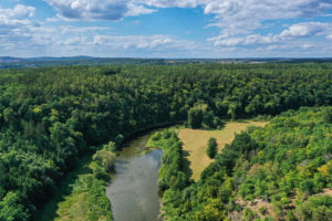 Pohled na lokalitu Háj z ptačí perspektivy. Lesnatý pás prochází podél ohybu koryta řeky od levé k pravé straně snímku. V popředí vlevo dole Zábělá – svah smíšeného lesa. Ve střední části řeka Berounka.