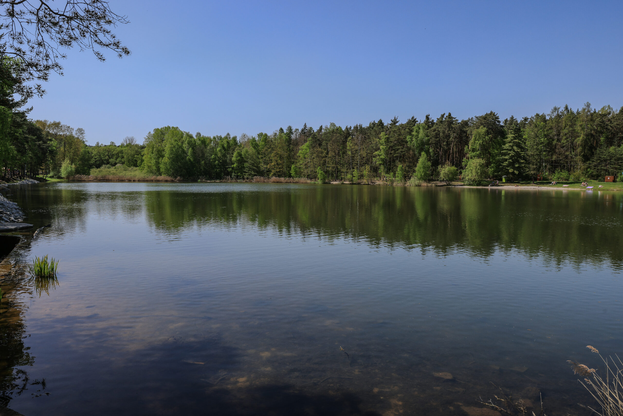 <h1>Šídlovský rybník</h1><br />
Na hladině průzračně čisté vody se odráží jehličnaté a listnaté druhy dřevin lemující okraj Šídlovského rybníka. V levé části fotografie se nachází pláž poskytující místo na vodní radovánky a odpočinek. 